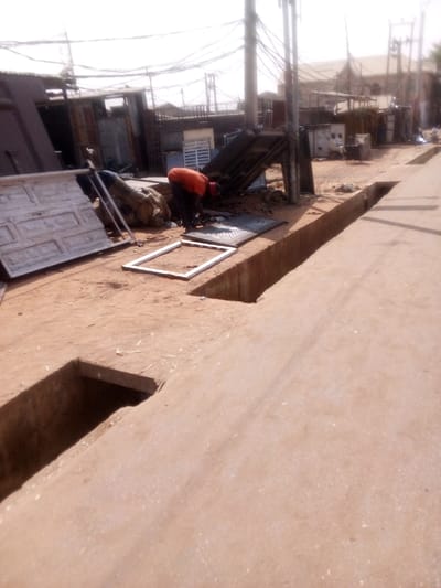 The scene depicts an outdoor workspace in Tudun Murtala, Nigeria. The ground is unpaved earth, with a concrete pathway and an open drainage trench extending along the right side of the frame. In the mid-ground, one individual, wearing an orange long-sleeved shirt, dark trousers, and a red head covering, is bent over, actively engaged with a dark, square, gridded object resting on the ground. A white, rectangular metal frame is positioned directly in front of this person. To the left, a large, rectangular white panel with embossed patterns leans against a stack of mixed materials, including what appears to be a light-colored, foldable ladder. The background features multiple utility poles supporting a dense network of overhead wires. Various low-rise buildings, some constructed with corrugated metal, are visible, suggesting a commercial or light industrial area. The scene is illuminated by strong natural light.