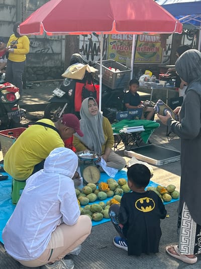 The image depicts an outdoor scene, likely a street market, in Makassar, Indonesia. A group of people are gathered around a stall covered by a red umbrella. In the foreground, a child wearing a black t-shirt with a yellow Batman logo is squatting. To the left, a person in a white hooded jacket is also squatting, facing away from the camera. Another individual, wearing a yellow t-shirt and a maroon baseball cap, is squatting and looking down at a scale. A woman in a grey hijab and mustard-colored shirt is seated behind the scale, appearing to be engaged in a transaction or discussion. Several piles of green mangoes are spread on a blue tarp. In the background, a motorcycle is parked, and there are signs with Indonesian text, including one that reads "BAKS UMUM Om Aldo." The ground is paved, and the environment appears to be urban.