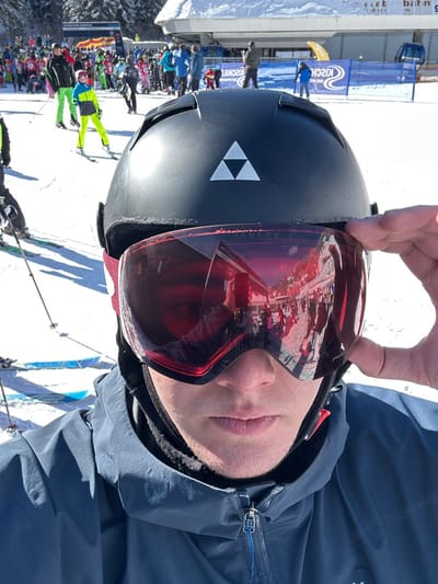The image displays a first-person perspective from a ski slope in Bad Hofgastein, Austria, under clear, sunny conditions. In the immediate foreground, an individual is visible from the upper chest to the top of the head, wearing a black ski helmet with a white Fischer logo (three inverted triangles) on the front. Red-tinted ski goggles are worn over the eyes, with the subject's right hand partially lifting the right side of the goggle frame. A dark blue ski jacket with a visible zipper and a black neck gaiter are also worn. The goggles reflect a crowded scene of skiers, snow, and distant structures.
The midground reveals a bustling, snow-covered ski slope populated by numerous skiers. Many individuals appear to be children or young adults, dressed in brightly colored ski apparel, including lime green and yellow suits. A blue banner, partially displaying the text "KISCHUL," is visible, indicating a ski school or event area. In the background, a large ski lift station featuring a snow-covered roof is present, alongside snow-covered mountains dotted with evergreen trees. The entire ground is uniformly covered in snow.