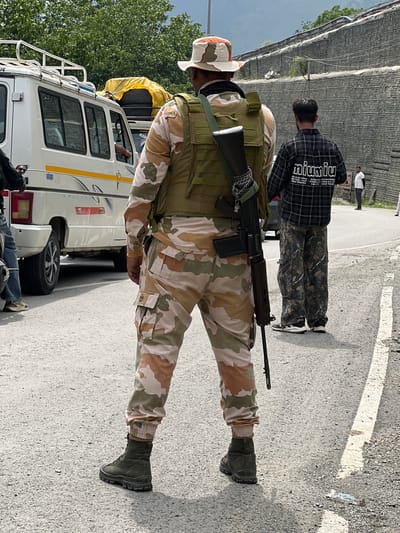In Gagangir, India, a person in camouflage uniform, wearing a tactical vest and a wide-brimmed hat, stands facing away from the camera. The uniform consists of a camouflage shirt and pants, and the person wears dark green boots. A rifle is slung across their chest, held by a chain. To the left, a white van with a roof rack is parked. Further down the road, another person wearing a plaid shirt and camouflage pants stands near a brick wall. The environment appears to be an outdoor setting with a paved road and trees in the background.