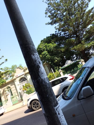 The image shows a street scene in Maputo, Mozambique, taken from a low angle, focusing on a large, grey lamppost, a white car, and a two-story, light-colored building. The car, a small hatchback, is parked facing the camera, with the driver's side partially visible. The car is parked in front of the building, which has a large, arched window, and the inscription "The CARD Company" and "Head Office" above it. Trees and foliage partially obscure the building, and there is another, darker car parked behind the white car. The scene is illuminated by the bright sun, casting long shadows.  The lamppost has white paint splattered on its surface, likely from a nearby construction project. The image appears to have been taken on a street in a residential area of Maputo, Mozambique. The latitude and longitude coordinates (-25.971, 32.575) confirm that the image was taken near a residential street in Maputo, Mozambique.