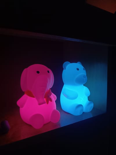 Two illuminated, cartoon-like figurines sit side-by-side on a dark shelf within a recessed space. The figurine on the left is pink, resembling an elephant wearing a scarf. The one on the right is blue and shaped like a bear. The background is dark, suggesting low ambient light. A small dark object is visible next to the pink elephant. The image location in the city of Cherkovitsa, Bulgaria, cannot be determined from the visual information provided.