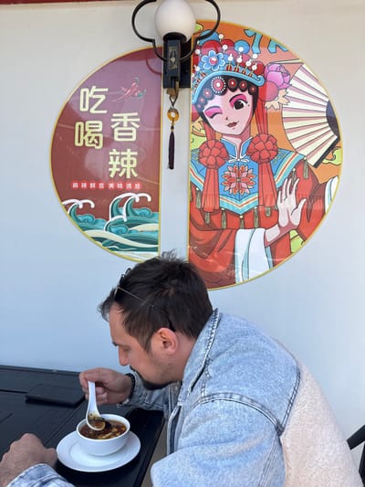 A man in a denim jacket with a sherpa collar is captured in a side profile, leaning over a dark table to eat soup from a white bowl. He uses a white spoon and has sunglasses pushed up onto his dark hair. The soup appears to be a dark, savory broth with various ingredients like mushrooms visible.
Behind him, on a light-colored wall, are two decorative semicircular panels, suggesting an Asian-themed eatery. The upper panel displays a vibrant, stylized illustration of a figure, likely a performer in traditional Chinese opera attire, adorned with an elaborate headdress and holding a fan. The artwork features a rich palette of red, orange, blue, pink, green, and yellow.
The lower panel, in a deep reddish-brown hue, includes Chinese characters and an illustration of a red chili pepper. The prominent characters read "吃喝" (chīhē), meaning "eat and drink," and "香辣" (xiānglà), meaning "fragrant and spicy." Below these, smaller text states "麻辣鲜香 美味诱惑" (málà xiānxiāng měiwèi yòuhuò), translating to "numbingly spicy, fresh and fragrant, delicious temptation." A decorative black light fixture with an amber-colored tassel hangs between the two wall art pieces.
The scene is brightly lit, indicating daytime. The man is engrossed in his meal, and the distinct decor points to a Chinese restaurant setting, located in Hainburg an der Donau, Austria.