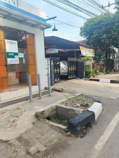 The image shows a street corner in Purwakarta, Indonesia (latitude: -6.551, longitude: 107.438). The street corner is situated between a bank building and a house with a black gate. The bank building has a sign that reads "BNI" and "ATM 24". There are four silver poles standing on the sidewalk in front of the bank building. The house has a sign that reads "PERCETAKAN RIZQ" and "Cetak". There is a partially obscured vehicle inside the gated area. The sidewalk is broken and cracked. There is a small drainage ditch next to the curb. The street is paved and there are white painted lines and black and white painted curbing. The sky is clear and blue. There are no people in the image.