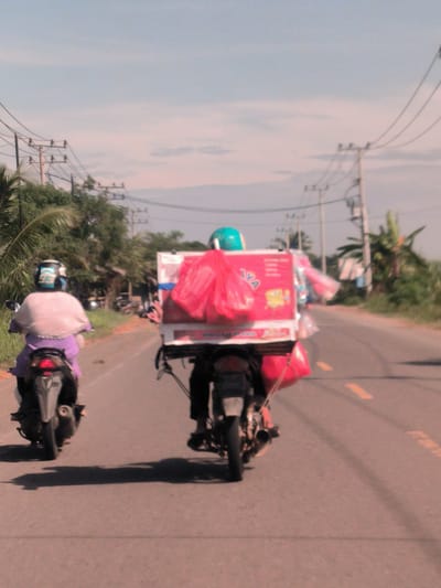 Two individuals are seen from behind, riding motorcycles down a paved road in what appears to be a rural or semi-rural area. The person on the left wears a white head covering and a purple outfit, riding a dark-colored motorcycle. The person on the right, wearing a turquoise helmet, is riding a motorcycle heavily laden with a large, rectangular, red delivery box on the back. Several red and pink plastic bags are attached to the box and motorcycle.

The road features a yellow dashed line in the middle and is bordered by lush green tropical vegetation, including palm trees. Numerous utility poles with power lines run along the side of the road and stretch across the sky. The sky is bright and hazy, with faint cloud formations. Strong shadows cast by the motorcycles and riders on the road indicate it is daytime and sunny.

On the prominent red delivery box, the letters "AYA" are clearly visible in white, possibly part of a brand name. Below this, there's a small yellow cartoon character and some indistinct white text, which seems to include "KUALITAS" (quality) or "TERBAIK" (best). The overall image has a slightly faded or reddish hue. The scene depicts everyday activity in a warm climate, possibly a vendor or delivery person at work.