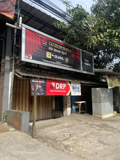 The scene is an exterior view of a store in Kecamatan Dayeuhkolot, Indonesia, located at latitude -6.971, longitude 107.617. The store has a large sign that reads "DIARUM SUPER #INI RISANYA SUPER 12 Rp 25.000,-," with additional text below it. Another sign is to the right of this one that reads "TOKO AZZARA." Below the large sign, there is a smaller banner advertising "DRP Djarum Retail Partnership" and "blibli mitra." There is a wooden door in the storefront, and a small, wooden table and stool are in front of it. The sidewalk in front of the store appears to be made of concrete and is cracked in several places. There are trees in the background and a few electrical wires overhead. No people are present.