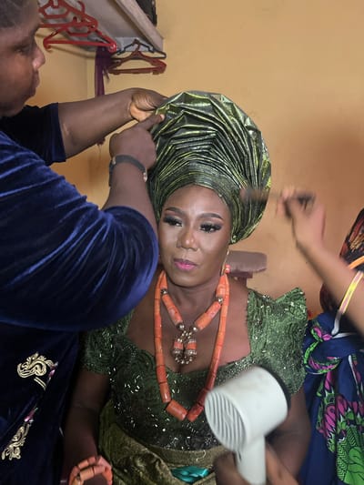 The image depicts a woman in Warri, Nigeria, undergoing preparations for an event. She is wearing a green, heavily embellished dress and a shimmering green headwrap. An orange beaded necklace adorns her neck. Two individuals are assisting her: one is positioning the headwrap, while the other uses a hair dryer near her chest. The environment appears to be an indoor space, possibly a dressing room, with clothes hangers visible in the background.