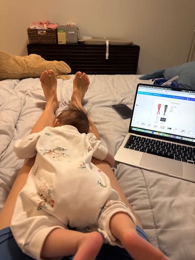 On a bed, a baby lies on a person's legs, facing away from the camera, wearing a white onesie with a koala print. Next to them is a laptop displaying a webpage with clothing items. A smartphone is visible above the baby's head, near the laptop. A brown wicker basket sits on a dark wooden shelf beside the bed, and a plush teddy bear is positioned near the person's feet. The bed is covered with a wrinkled light grey sheet.