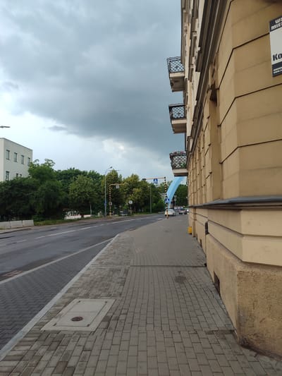 The image captures a street scene in Tczew, Poland, on an overcast day. On the right is the side of a light yellow building with visible architectural details such as cornices, decorative elements, and balconies with wrought-iron railings. A sign is partially visible on the building's facade. A sidewalk paved with gray rectangular bricks runs along the building. To the left of the sidewalk is a street with two lanes of asphalt. A pedestrian crossing with a traffic light is visible further down the street, accompanied by trees and other buildings in the background. A blue archway structure spans over the road, and several cars are present on the street.