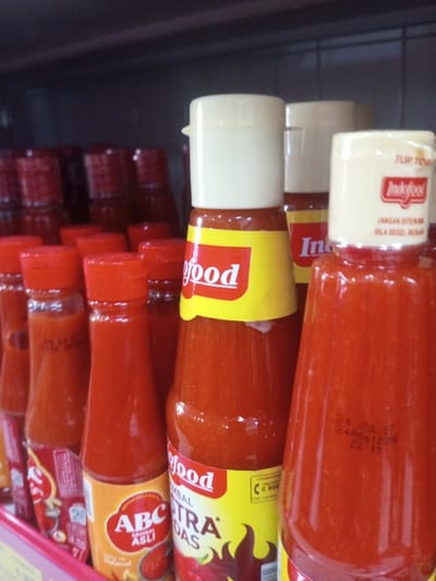 A close-up image taken in a retail environment, likely a supermarket or grocery store, in Naung Bo Oe, Myanmar, depicts multiple bottles of chili sauce arranged on a shelf. The foreground prominently features a tall, clear glass bottle of Indofood chili sauce with a cream-colored flip-top cap, displaying a visible expiration date of "24 JUL 27" and batch code "24R6612516" printed directly on the glass. The cap also shows the "Indofood" logo and text "JANGAN DITERIMA BILA SEGEL RUSAK" (Do not accept if seal is damaged). To its left, another Indofood bottle with a yellow label for "Indofood Sambal Ekstra Pedas" (Extra Spicy Chili Sauce) is visible, also topped with a cream-colored flip-top cap. Further left, an "ABC Sambal Asli" (Original Chili Sauce) bottle with an orange label and a red flip-top cap stands. Numerous other bottles, predominantly red with red caps, are visible in the blurred background on a dark shelf, extending into the upper left of the frame. All bottles contain a reddish-orange viscous liquid. No individuals are present in the scene.