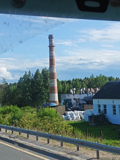 The image shows an industrial facility on a clear, sunny day, with a bright blue sky and scattered white clouds. In the foreground, a metal guardrail runs along the edge of a road. Beyond the guardrail, a large, red and white striped smokestack dominates the scene, its brickwork weathered and discolored. The smokestack is surrounded by lush green trees and vegetation. To the right of the smokestack, a cluster of industrial buildings can be seen. One building is painted white with blue trim and has several windows. Another building, to the left, appears to be made of concrete and has a dark roof. Piles of white sacks are stacked near the buildings. The overall impression is one of an active, though perhaps older, industrial site set against a natural backdrop. The specific location is an industrial zone in the city of Torzhok, Russia.