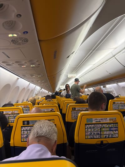 The image shows the interior of a Ryanair airplane cabin, possibly taken during a flight or while stationary on the ground, as indicated by the Ryanair branding on the overhead compartment and seat headrests. Passengers are seated in rows with yellow headrests that have safety instructions. The aisle extends into the distance, with more passengers seated further down the cabin. Some passengers are interacting or moving around. The environment appears to be within a commercial airplane cabin, with overhead lights and air vents visible. The image does not provide evidence of the location being in Luqa, Malta.