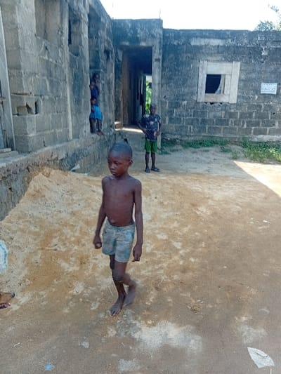 The image captures an outdoor scene featuring several children and stone buildings under natural daylight.
In the foreground, slightly to the right of the center, a young boy stands facing generally towards the viewer. He appears to be around 5-10 years old, with dark skin and very short dark hair. He is shirtless and wears light-colored, possibly patterned, shorts. His expression is neutral. His bare feet are visible on the sandy ground.
To the left and in the mid-ground, a series of buildings constructed from large, rough-hewn stone or concrete blocks stretch across the frame. These structures appear somewhat weathered or unfinished. One section of the building, further back, features a rectangular window opening with a dark interior visible through it. Above this window, high on the wall, there is a small, light-colored rectangular plaque or sign with some faintly visible, unreadable text. Another section of the building has a large, open doorway, through which a dark interior is also visible.
Standing in or near this large doorway, further back than the main foreground boy, is another child or young person. They are wearing a dark, short-sleeved shirt with a light pattern and light-colored shorts. Their back is mostly towards the viewer, making their face unclear. Even further back and to the left, partially obscured by the buildings, a third smaller child can be seen, wearing a blue top and lighter bottoms, appearing to stand or sit low near the base of the building.
The ground throughout the scene is uneven and composed of light brown and tan dirt or sand, showing various textures and small patches of debris. A small, crumpled white object is visible on the ground near the feet of the foreground boy. The lighting is bright and natural, indicating it is daytime, though the absence of strong shadows could suggest either an overcast day or the sun being high in the sky. The overall atmosphere is candid, depicting children in what appears to be their everyday environment.