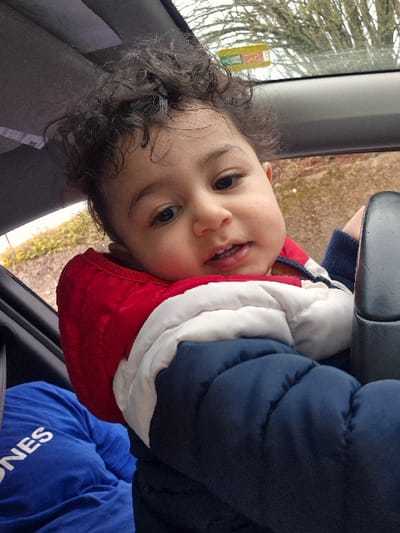 The image captures a young child seated inside a vehicle, located in Melle, Belgium. The child, with dark curly hair and dark eyes, is positioned prominently in the foreground, facing the camera with a slight open-mouthed smile revealing small teeth. They are wearing a puffer jacket with a distinct tri-color pattern: red at the top, a white band across the middle, and dark blue covering the lower torso and sleeves. To the child's left, a person wearing a royal blue t-shirt with white partial lettering "ONES" is partially visible. The vehicle's interior features a light gray headliner and a dark gray vertical element on the right side of the frame. Through the vehicle's rear window, a blurred outdoor environment is visible, comprising bare deciduous tree branches against an overcast sky and brown ground. A rectangular sticker, yellow with red design elements, is affixed to the upper right portion of the rear window.