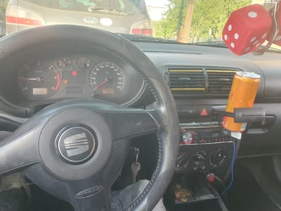 The image provides a close-up view of the interior of a car, focusing on the dashboard and steering wheel. The driver's perspective is evident, with the steering wheel prominently featured in the foreground, displaying the "SEAT" logo. Keys are hanging from the ignition.

To the left of the steering wheel, the car's instrument cluster is visible. The speedometer reads a speed in kilometers per hour, and the odometer shows a mileage of 305926. A digital clock displays "19:17", suggesting it is late afternoon or early evening. The tachometer, also part of the instrument cluster, indicates engine RPM.

To the right of the steering wheel, the dashboard features an air conditioning vent with gold-colored slats. A can of an orange beverage is placed in a cup holder. Above the vent, a pair of fuzzy red dice, a common car accessory, hangs from the rearview mirror. The car's stereo system, a "Pioneer" brand, is also visible, with a display showing "12:00". Various control knobs and buttons for the car's climate control and audio system are present.

Outside the car, through the windshield, trees and foliage suggest an outdoor setting, possibly a street or parking area. The lighting indicates it is daytime, with the sun casting shadows. The overall scene depicts a mundane moment inside a parked or stopped vehicle. The visible text includes "SEAT", "AIRBAG", "rpm x 1000", "km/h", "Pioneer", and "19:17" on the dashboard displays, along with what appears to be a model number or indicator "531.1" on the odometer. The location context indicates Bulgaria.