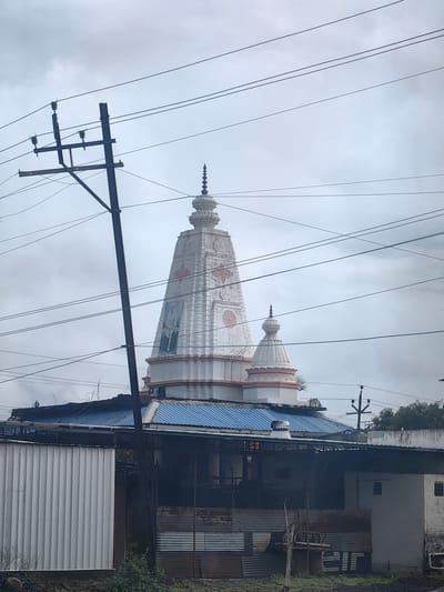 This image depicts a temple in Mohegaon, India, with a prominent white spire adorned with decorative patterns and a portrait of a person. The temple is partially obscured by a power pole with electrical wires stretching across the frame. In the foreground, there's a low-rise structure with a blue corrugated roof and walls made of corrugated metal and white siding. The background shows a cloudy sky.