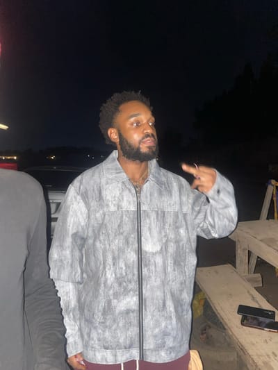 A person with dark skin, a beard, and dark curly hair is wearing a gray jacket and black pants. The person is giving a hand gesture. Two phones are visible on a wooden surface above the person. The location is outdoors at night, possibly in Galadimawa, Nigeria.