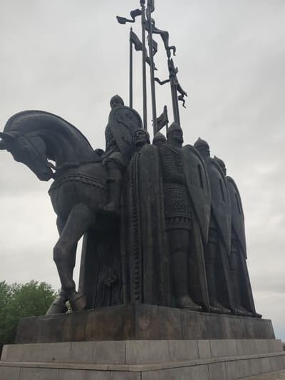 The image depicts a bronze monument in Khotitsey, Russia. The monument consists of a large, sculpted grouping of medieval figures, including a horse-mounted warrior at the front. Above and behind him, several other figures stand in a row, holding shields and spears with banners attached. The background is a cloudy sky. The monument sits on a large concrete plinth, with some greenery visible at the bottom right of the frame.