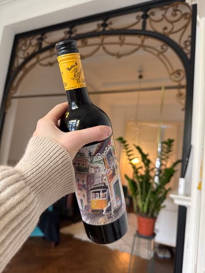A light-skinned hand, adorned in a light beige ribbed long-sleeved sweater, holds a dark glass bottle of "Porta 6" red wine. The bottle features a colorful label depicting a Lisbon street scene with traditional buildings, laundry, and a yellow tram. The bottle's neck is sealed with a black screw cap and a yellow foil label displaying "Porta 6" and a cartoon character playing a guitar. The image is captured facing into a large mirror, which reflects an interior living space. The mirror itself has an ornate, dark metal arched frame with gold-colored scrollwork details at its apex. Reflected in the mirror, to the right of the bottle, is a large green potted plant in an orange terracotta-colored pot, positioned on a light-colored stand. Further into the reflection, parts of a light-colored wall, a blurry light fixture (possibly a chandelier), and a white fireplace mantelpiece are visible. The floor reflected appears to be a warm brown, likely wood. The specific location within Saint-Gilles - Sint-Gillis, Belgium, is not ascertainable from the depicted indoor scene.