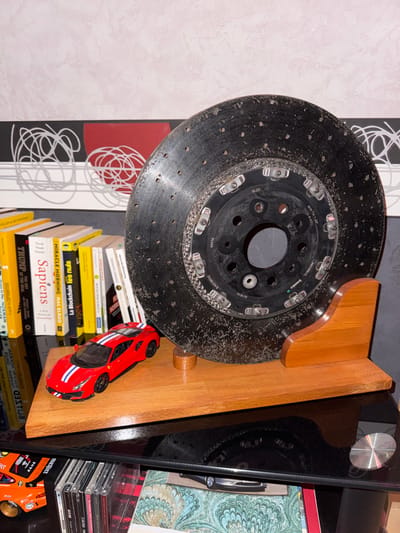 A brake rotor is mounted on a wooden base and displayed on a shelf amidst books, CDs, and decorative items. The rotor, visibly used and worn, adds an industrial element to the space. The books, including "Sapiens" and "Miracle Morning," suggest an eclectic reading taste. A red toy car rests on top of the book stack, providing a pop of color and a playful contrast to the serious tone of the other objects.