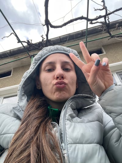 A woman is outdoors, wearing a grey puffer jacket with the hood up. She is making a peace sign with her left hand and puckering her lips. She has brown hair. In the background, there is a building with a window and a bare vine growing along the structure. Based on the location provided, this scene is in Hrishteni, Bulgaria.