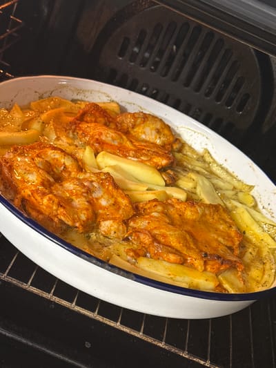 A white oval baking dish with a blue rim is filled with chicken and potato wedges. The chicken pieces are seasoned with spices, giving them a reddish-orange hue and appear to be cooked to a tender state. The potato wedges are golden brown and interspersed with the chicken. The baking dish is placed on a rack inside an oven, with the oven's heating element visible in the background. The image is likely taken in a kitchen in Rogosh, Bulgaria.