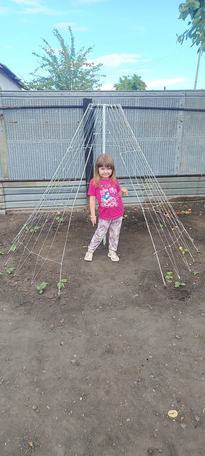 A young girl stands inside a conical trellis structure in a garden. She is wearing a pink t-shirt with a graphic print of roller skaters and a unicorn, and matching pink pants with a unicorn pattern. Her left hand is extended downwards, pointing at the ground. The trellis is made of white strings forming a net-like structure, supported by poles. Several small green plants are visible in the dark soil around the base of the trellis. In the background, there is a corrugated metal fence and some trees under a partly cloudy blue sky. The location is the city of Khokhly, Russia.