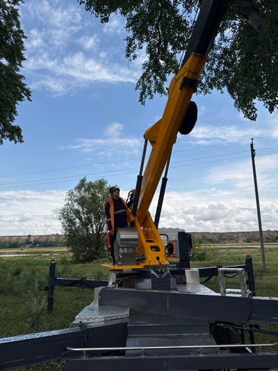 A man wearing a high-visibility vest and a hard hat stands on a yellow crane. The crane is positioned on the ground in a grassy area. In the background, there is a body of water, a hill with trees, and a power line. The sky is blue with some white clouds.