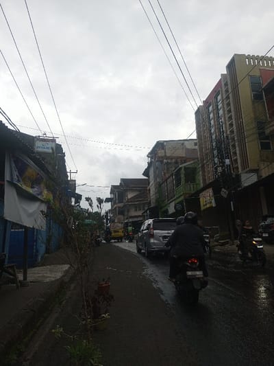 The image depicts an urban street scene in Sindanglaya, Indonesia, under an overcast sky. The asphalt road, wet from recent precipitation, extends into the background, flanked by buildings on both sides. In the immediate foreground, a dark-colored motorcycle, license plate F 3243 YO, is positioned with its rear visible, carrying two individuals wearing helmets. Ahead of the motorcycle, a silver SUV is parked or moving slowly. Further down the street, a yellow van and additional motorcycles are visible.
On the left, a concrete sidewalk borders the road, displaying potted plants and thin, leafless shrubs. Buildings with corrugated roofs and visible signs, including "PERJAPAK" and "INMARCH," line this side. A building with a blue lower section is also present. On the right, taller, multi-story buildings stand, one featuring a distinct yellow-beige facade with vertical red-brown accents and multiple windows. Numerous black electrical and communication cables are strung overhead, crisscrossing the upper portion of the frame. On the right sidewalk, two women are visible, one in a dark dress, and another motorcycle is parked or moving. The overall environment suggests a busy commercial or residential street.