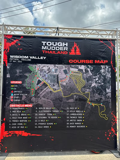 The image shows a course map for "Tough Mudder Thailand" at Wisdom Valley, Pattaya, Thailand, scheduled for May 2nd, 2026. The map is mounted on a metal frame outdoors, and the sky is visible in the background.
The map details the course layout for both 15K and 5K races, including the start and finish lines, obstacle locations, and water stations. A legend describes the symbols used to represent these elements.
A "Obstacle Menu" lists 21 obstacles, including "Hydrophobia," "Mud Mile," "Devil's Bread," "Berlin Walls," "Electroshock Therapy," and "Monkey Business," each marked with either one or two dots.