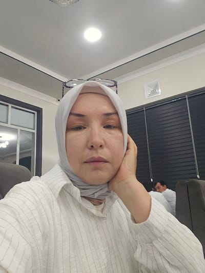 A woman wearing a light gray hijab and a white pinstriped button-down shirt is shown in a close-up, frontal shot. She is looking down and slightly to her left, with her right hand resting on her cheek. Her eyeglasses are perched on top of her head. In the background, another person wearing a white shirt is visible. The setting appears to be an indoor space with white walls, a drop ceiling with recessed lighting, and dark blinds on a window. The image was likely taken in Nurafshan (Toytepa), Uzbekistan.