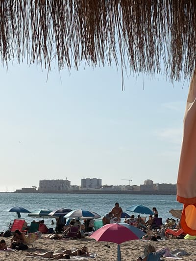 This image depicts a sunny day at a beach, likely La Caleta beach in Cádiz, Spain, given the distinctive architecture of the buildings in the background which are characteristic of the city. In the foreground, the scene is framed by the edge of a thatched umbrella and a draped orange fabric. The sand is populated with numerous beachgoers relaxing under various umbrellas, some in beach chairs, others lying on towels. The sea is visible in the middle ground, with several people wading or swimming. A distant cityscape with white buildings and a construction crane marks the urban background. The sky is clear and pale blue, suggesting a warm, bright day.