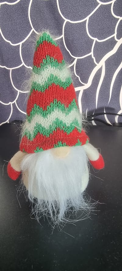 This is a close-up, horizontal shot of a gnome decoration. The gnome is positioned on its side, with its pointed hat extending towards the left of the frame and its fluffy white beard filling the right side. The hat is knitted in a zigzag pattern of red, green, and white stripes, and is trimmed with white fluffy material. The gnome has a small, round nose and red shoe-like feet. The background on the left is a dark blue fabric with a white abstract pattern. The background on the right is a dark, textured surface, possibly a table. The lighting suggests it is daytime, but there are no clear indicators of the time of day or weather. There is no visible text in the image. The setting appears to be indoors, possibly a home environment in Naaldwijk, Netherlands. The overall impression is that of a festive or decorative item.