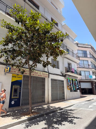 This media file captures a street scene in Sant Antoni De Portmany, Spain. The image is taken from a low angle, looking up at a multi-story building with a tree in the foreground. The building appears to be old, with various shops and businesses on the ground floor. One shop has a sign that reads "Kaldict idad R. Torres CLUB - ALTA FIDELIDAD Y FOTOGRAFIA" and another sign for "LECTRIC SHOP VIDEO VI". An ATM machine, labeled "ATM - Cajero" and "Euronet. ING", is also visible. Above the shops, there are windows with security bars, and laundry is hanging from some of the balconies.

In the lower right portion of the image, a woman wearing a bikini top and denim shorts is partially visible, bent over. The scene appears to be set during the daytime, with bright sunlight casting distinct tree shadows on the pavement, suggesting a sunny day. The architecture and the presence of an ATM machine indicate an urban setting. The overall impression is a bustling street in a coastal town, likely during the warmer months given the woman's attire.