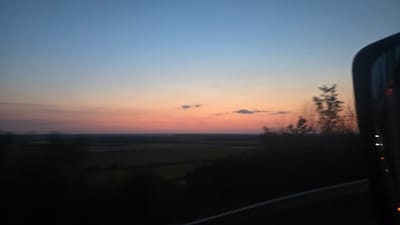 The image depicts a rural landscape at dusk. The sky transitions from a light blue at the top to pink and orange hues near the horizon, suggesting a sunset or sunrise. Scattered clouds are visible in the sky. In the foreground, the dark silhouettes of trees and vegetation obscure the ground. To the right, a portion of a vehicle's side mirror is visible, reflecting some lights. The overall impression is one of a peaceful, natural scene viewed from a moving vehicle. The location is identified as Pleshivets, Bulgaria.