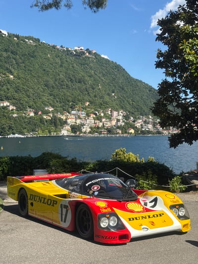 The image captures two Porsche 962 racing cars, parked on a roadside with a scenic view of Lake Como, Italy, in the background. The cars are decorated with Shell and Dunlop branding. The hillside across the lake is visible, dotted with buildings characteristic of the region. The sky is clear, and there's foliage in the foreground, suggesting a tree or shrubs. The overall scene suggests an outdoor setting on a sunny day in the city of Como, Italy.