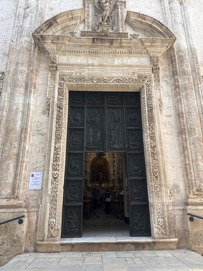 The image presents a grand, dark-colored door set into a weathered, light-stone facade, belonging to a historic religious building in Monopoli, Italy. The door, likely made of wood or metal, is composed of numerous rectangular panels, each featuring a carved relief. Notably, one panel prominently displays a full skeleton, while others depict skulls, crossbones, and tools such as a pickaxe, shovel, and scythe, serving as memento mori symbols.

The intricate stone facade surrounding the door is adorned with elaborate floral motifs, scrollwork, and a frieze above the entrance that also incorporates skull and crossbone imagery. A Latin inscription, partially visible on the left vertical frame of the door, reads "QATIO.PRO.DEFVNC.TIS.EXORA.R," suggesting prayers for the deceased.

The door is partially open, revealing a glimpse into the opulent interior of what appears to be a church or cathedral. A large, gilded altarpiece or artwork is visible deep inside, and several indistinct figures of people can be seen, indicating ongoing activity within the building.

Below the door, a small white printed sign is affixed to the stone wall. The word "MONOPOLI" is discernible at the top, along with "Cattedrale della... Monopoli," confirming the location and type of structure. The sign also features a small photograph of a person and Italian text, likely an announcement or schedule. The scene is illuminated by natural daylight, suggesting a clear day. The overall impression is one of historical grandeur and solemnity.