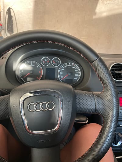 This is a close-up, eye-level interior shot of a car, likely taken from the driver's seat. The focus is on the steering wheel and dashboard of an Audi vehicle. The steering wheel is black leather with red stitching and perforated sections on the grips. The Audi logo, four interlocking rings, is prominently displayed in the center of the steering wheel, outlined in chrome. Below the logo, the word "AIRBAG" is embossed in the leather.
The dashboard features two main gauges: a speedometer marked up to 260 km/h and a tachometer marked up to 6000 RPM (labeled "x 1000"). Smaller auxiliary gauges are also visible, including what appears to be a fuel gauge and a temperature gauge. To the right, a section of the air vent is visible, as is a red indicator light on the center console.
The background is blurred, showing a beige, textured wall, possibly the interior of a garage or a building. The lighting suggests it is daytime, with sunlight casting shadows and highlights. The lower part of the image shows the thighs of a person, indicating the vantage point is from someone seated in the driver's seat. The scene depicts a static interior of a parked vehicle.
There is no information within the image that allows for the determination of the specific location being Rogosh, Bulgaria.