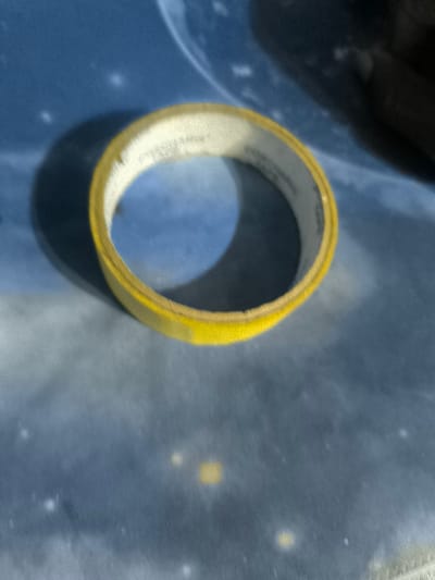 The image shows a roll of yellow tape lying on a blue surface. The tape is partially unrolled, revealing its yellow adhesive side. The surface is textured and appears to be made of fabric. There are no people, objects, or actions present. The coordinates provided are for the city of Sapele, Nigeria, but the image does not provide any context for the location. There are no indications that this image was taken in Sapele, Nigeria.