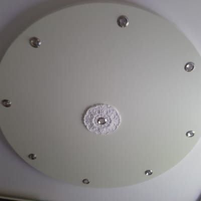 The image shows a close-up, overhead view of a round, white ceiling fixture with several small, recessed lights. In the center of the fixture, there is a decorative white rosette with a silver-toned embellishment in its middle. Eight small, circular lights are evenly spaced around the perimeter of the fixture. The image appears to be taken indoors, likely in a residential or commercial setting. There is no information in the image to suggest a specific location in Sambir, Ukraine.