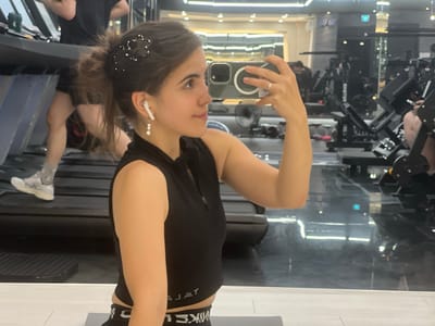 This is a media file capturing a woman taking a selfie in a gym. The woman has dark hair pulled back in a ponytail, adorned with a flower-shaped hair clip. She is wearing a black sleeveless top and black workout bottoms with visible white lettering that appears to read "NIKE". She has white wireless earbuds in her ears and is wearing a ring on her finger.
The setting is a gym with treadmills visible in the background, along with various weight machines. The floor is reflective, suggesting it might be a polished surface or a mirror. Other people can be seen in the background, engaged in their workouts. The lighting appears to be artificial, consistent with an indoor gym environment. The overall mood is one of fitness and personal activity. There are no clear indicators of the time of day or weather conditions. The location context is Daegu, South Korea.