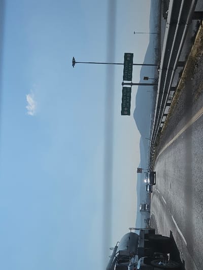 A highway scene with a tanker truck on the right. The road curves ahead with other vehicles in the distance. Roadside barriers line the left. A highway sign directs drivers to Queretaro/Celaya and Ciudad de Mexico/San Juan del Rio. Mountains are visible in the background under a blue sky with a small cloud.