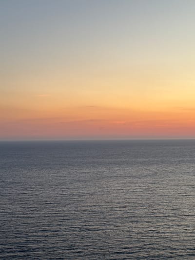 The image depicts a serene seascape at sunset. The sky transitions from a pale blue at the top to a warm gradient of orange and pink near the horizon. A thin band of clouds is visible in the distance, catching the sunset colors. Below the sky lies the calm sea, its surface rippled with gentle waves reflecting the muted tones of the sky. The water is a deep, muted blue-gray. The scene is devoid of any human presence, structures, or distinctive landmarks, suggesting an unobstructed view of the natural environment. The overall impression is one of tranquility and natural beauty, characteristic of coastal landscapes. The location in Glavatičići, Montenegro, implies a possible view of the Adriatic Sea.