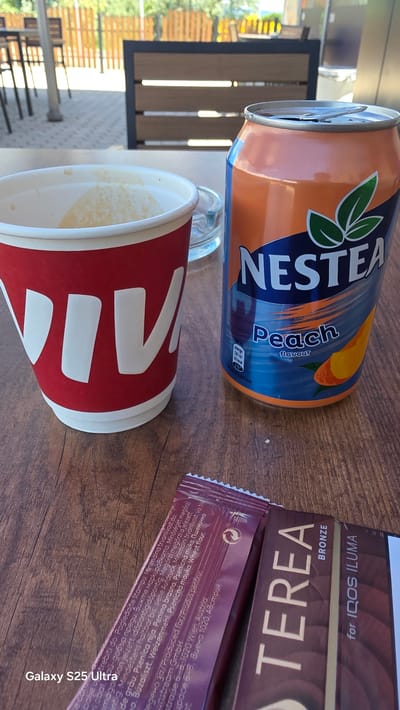 An outdoor table setting in Kaspichan, Bulgaria, features a disposable coffee cup with a red and white design, partially filled with a frothy beverage. To its right, an orange and blue can of Nestea Peach flavored iced tea is visible, with the lid open. In the foreground, two packages are laid out on a wooden table: one is a dark purple, foil-wrapped item with white text that includes "Galaxy S25 Ultra" in the lower left corner and additional text in multiple languages. The other package, to the right, is a brown box with the word "TEREA" in large, gold letters and "BRONZE" printed below it, along with "for IQOS ILUMA". The background shows a blurred view of outdoor seating and a wooden fence.