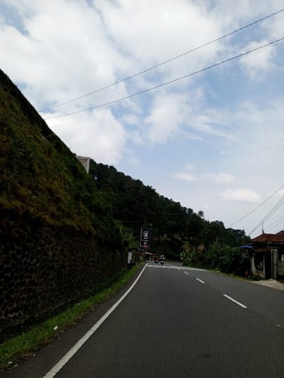 The image depicts a rural or semi-rural road scene in Ciloto, Indonesia, under a partly cloudy sky. The asphalt road occupies the lower-center of the frame, featuring a solid white line marking its left edge and dashed white lines indicating lanes further ahead. To the left, a steep, dark green, vegetation-covered hillside rises abruptly, supported at its base by a dark stone retaining wall. Utility wires traverse the upper portion of the sky. Along the right side of the road, dense green foliage obscures several modest, weathered structures. A large black banner with white text and a logo hangs vertically near these structures. Two motorcycles, one red and one lighter colored, are visible on the road in the mid-ground, heading away from the viewer or stationary. Small figures, likely people, are discernible near the motorcycles and structures on the right. The overall environment suggests a mountainous or hilly region with local residences alongside a main thoroughfare.