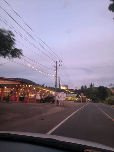 This image depicts a roadside scene at dusk in what appears to be a rural or semi-rural area of Sukamulia, Indonesia. The sky above is a soft blend of purple and pink, indicating twilight, while artificial lights from businesses along the road have already been switched on.

On the left side of the asphalt road, several buildings, likely restaurants or cafes, are brightly illuminated with numerous string lights, casting a warm glow. These establishments feature red-orange and white elements in their architecture. A black car is parked in front of one of these buildings. Beyond the immediate structures, more lights and buildings can be seen, leading up to a backdrop of densely forested hills. Utility poles and a dense network of power lines dominate the left and center-mid sections of the sky, running alongside the road.

A prominent signpost near the road displays text. The largest text clearly reads "PAPI KOPI ACEH BARU" (Papi Coffee New Aceh), with smaller text beneath it stating "Mie Ungket Saree & Nasi" (Saree Squid Noodles & Rice). This signage points to local Indonesian food and coffee, aligning with the provided location context of Aceh.

The road itself is well-marked with white and yellow lines. The perspective suggests the photo was taken from inside a vehicle, with part of the dashboard and windshield visible in the foreground. A blurred tree branch in the upper left corner implies movement. While no specific individuals are visible, the illuminated businesses and parked car indicate human activity, suggesting the scene is of a functional community either winding down or opening for evening trade. The atmosphere is peaceful, capturing the transition from day to night.
