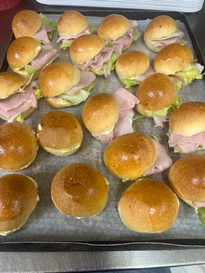 This media file shows a tray of partially assembled sandwiches, likely mini ham and lettuce rolls, resting on a baking sheet lined with parchment paper. The sandwiches are arranged in neat rows, with some fully closed and others open, revealing layers of pink ham and crisp green lettuce, along with a white spread that appears to be mayonnaise. The buns are golden brown and appear to have been recently baked, with a slight sheen on their surface. Some buns are whole, while others are cut in half. The baking sheet is placed on a metallic surface, possibly a countertop or a part of a kitchen appliance, and a dark, ribbed edge of a baking tray is visible along the right side of the frame. The setting appears to be a home kitchen, given the informal arrangement of the food and the presence of what looks like a baking tray. The lighting suggests it's daytime. No people are visible in the frame. The location context of Vallo della Lucania, Italy, suggests a domestic preparation of food, possibly for a picnic or a casual meal.