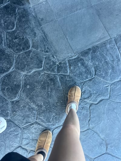 A top-down shot taken in Jesús del Monte, Mexico, shows a section of dark grey, patterned stone pavement. Two female legs are visible; one is angled towards the upper right with a light-colored sock and a tan slipper, while the other extends diagonally from the center to the lower right, wearing a darker sock and tan slipper. A glimpse of what appears to be a white disposable cup/container is located towards the bottom of the frame. The pavers are of irregular shapes, separated by darker indented lines.