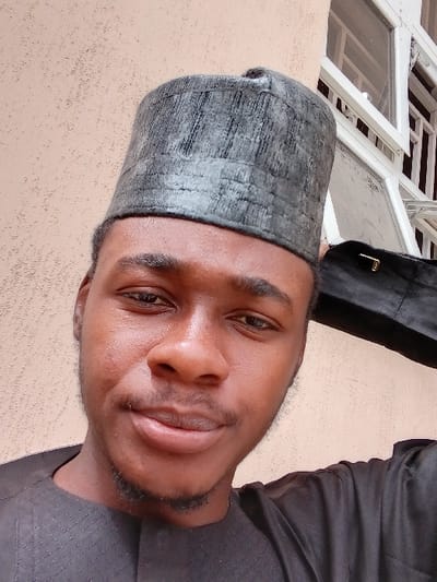 A male subject, appearing to be a young adult, is depicted in a close-up, chest-up photograph taken outdoors in the city of Kwazailewa, Nigeria. He has dark skin, short dark hair, and a visible beard and mustache. He wears a dark, textured traditional cap and a dark garment, possibly a traditional top, visible at the shoulders and neck. The subject is looking directly at the camera with a neutral to slight smile. The background on the left consists of a textured, light beige or tan building wall. On the right, a white-framed window with multiple panes is partially visible, behind which a darker, possibly black, object with a metallic clasp is present over the subject's right shoulder.