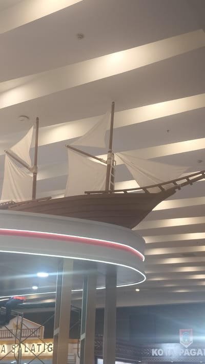 The image depicts a decorative model of a ship with white sails, suspended from a ceiling. The ceiling features a modern design with recessed lighting strips. Below the ship, there is a curved architectural element with red and white lighting. In the lower part of the image, scaffolding is visible, suggesting ongoing construction or maintenance. A person wearing an orange vest is working on the scaffolding. Text on a banner in the lower left reads "A SALATIG". In the lower right, a crest and the text "KOTA PAGAI" are visible. The lighting suggests it is daytime, and the indoor setting indicates a public space like a mall or convention center in the city of Medan, Indonesia.